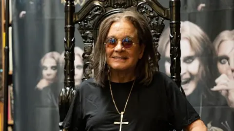 Getty Images Ozzy Osbourne - a man with long dark hair, sunglasses, a necklace with a crucifix attached, and a black shirt, sits on a black gothic-style throne and smiles. Behind him is a large banner featuring multiple images of him.