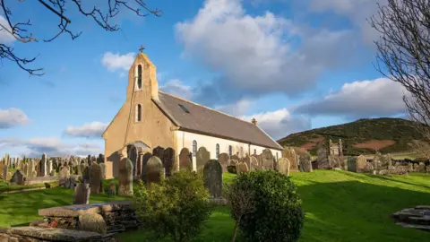 Manx scenes A hill-top view of Kirk Maughold church, dedicated to the saint, surrounded by gravestones on green grass banks, with a hill in the background