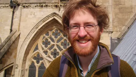 Michael Manning Michael Manning smiles, he has a ginger beard and hair and wears small spectacles. He wears a a jumper and a backpack and stands in front of a church that has stained glass windows.