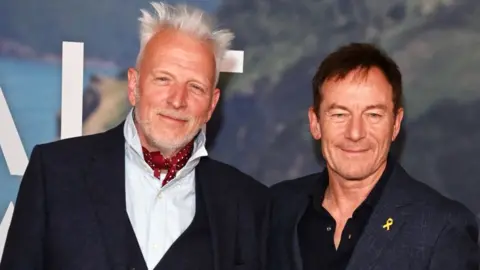 Getty Images Moth Winn (L) and Jason Isaacs attend the UK special screening of The Salt Path at The Curzon Soho in London. Winn is wearing a dark suit, white shirt and a red and white spotty cravat. Isaacs is wearing a black suit and open-neck shift.