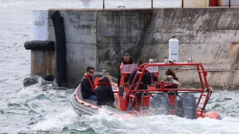 Peter Jolly/Northpix Tom Holland, wearing sunglasses and a red life jacket over a dark jacket, is sitting in an inflatable boat powered by two large outboard engines. There are at least five other people with him - three women and two men. The boat's engines are causing the water to foam as they head for the harbour entrance.