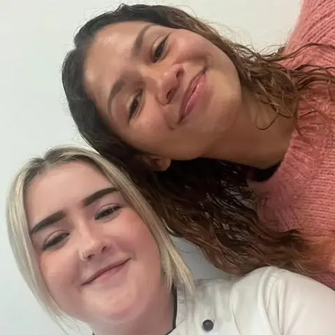 Miele's Gelateria Zendya has brown wavy hair, a pink jumper and is smiling as she poses for a selfie with Joss. Joss has blonde hair and is wearing an ice cream shop outfit.