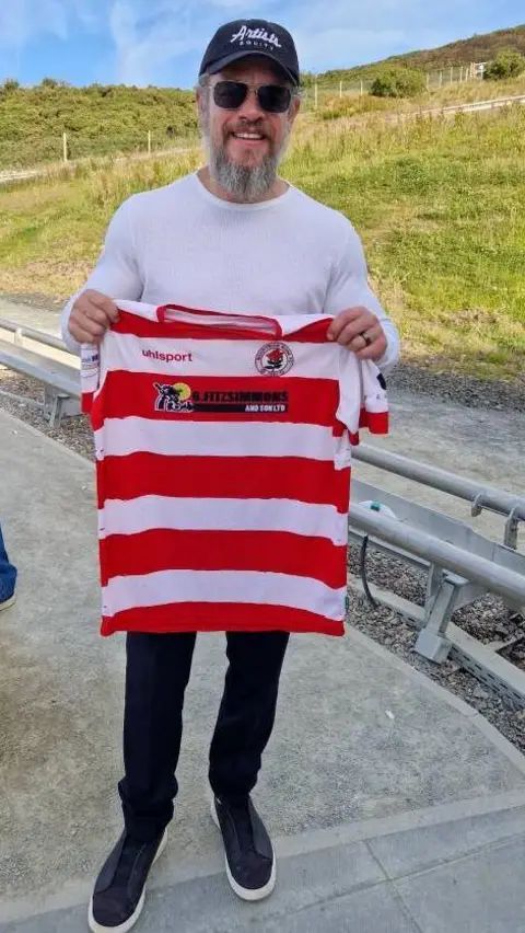 Bonnyrigg Rose Matt Damon is wearing a baseball cap, a pair of shades and a white long-sleeved top. He has blue jeans and trainers. He is standing outside and is holding a red and white striped Bonnyrigg Rose top.