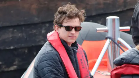 Peter Jolly/Northpix A close up of Tom Holland, wearing sunglasses and a red life jacket over a dark jacket, is sitting in an inflatable boat. He has his brown hair in his trademark tousled style. Behind him is a grey harbour wall. 