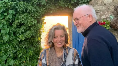 Sue Butler Sue Butler, with blue eyes and grey hair and a loose white, blue and orange poncho style top, with her husband Eddie Butler, a tall man with grey hair and glasses and a navy blue turtle neck sweater. They're stood in front of a stone house with an open blue door and a wall of green leaves 