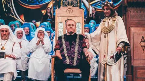 Eisteddfod Genedlaethol Cymru A man with short ginger hair and beard wearing a purple waistcoat with gold embroidery. He is sat in a wooden chair and is surround by people wearing white and blue robes. Stood next to his chair is a woman wearing intricate gold robes, a large gold necklace and crown.