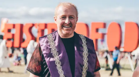 Eisteddfod Genedlaethol Cymru A man with short ginger hair and beard, stood in front of large red lettering forming the word Eisteddfod. He is wearing an orange and black t shirt and purple waistcoat with gold embroidery, and is smiling while looking at the camera.