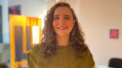 BBC Caoimhe Ní Cathail is looking at the camera and smiling. She has long, curly brown hair and is wearing a large gold earring in her left ear. She is wearing a moss green top. 