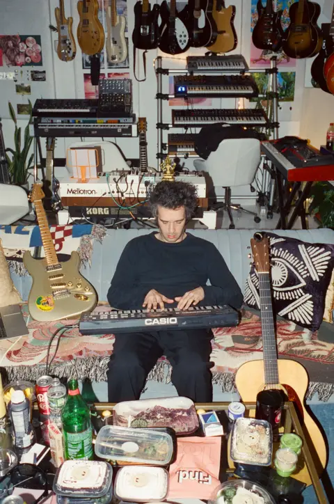 Jordan Curtis Hughes Matty Healy from The 1975 plays a keyboard while surrounded by clutter and musical instruments in what looks like a living room 