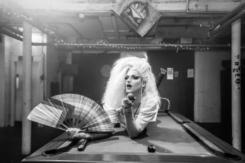 Jen Amelia Veitch Image of a clubber with long, blonde hair holding a fan and a billiard ball, lying on a billiards table