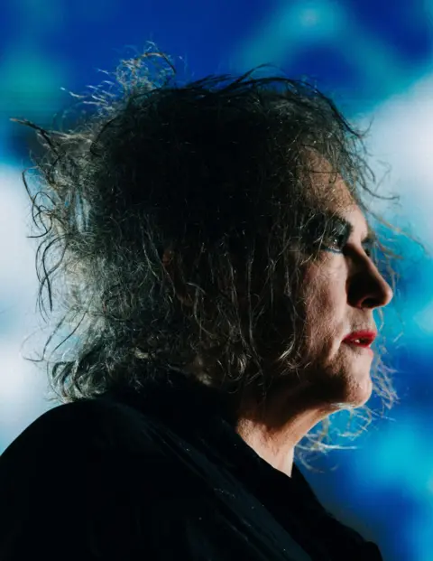 Phoebe Fox Profile shot of The Cure's Robert Smith against a blue background. Smith is wearing black eye make up and red lipstick.