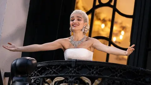 Jeff Spicer/Getty Images Actor Rachel Zegler performing Don't Cry For Me Argentina live on the balcony as she plays Eva Peron in Evita at the London Palladium. She has blonde hair in an elegant, low braided bun and it wearing a strapless white dress and an ornate diamond necklace. Her arms are stretched out and she is smiling while singing.