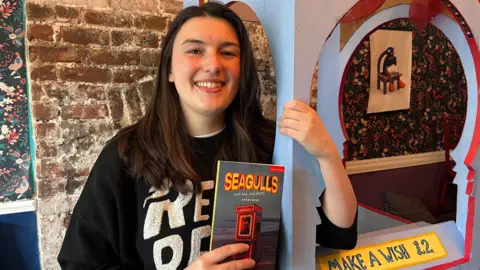 BBC Sarah Bond, 28, is stood holding part of the set used in Seagulls and Sad,  Sad Stories while holding a published manuscript of the play. She is dressed in a black hooded jumper which has white writing across it which reads "Rebel, rebel".