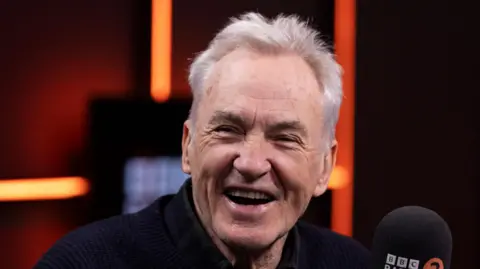 Jamie Simonds/BBC Larry Lamb, a man smiling at the camera. He is sitting next to a microphone.
He is wearing a black top. 