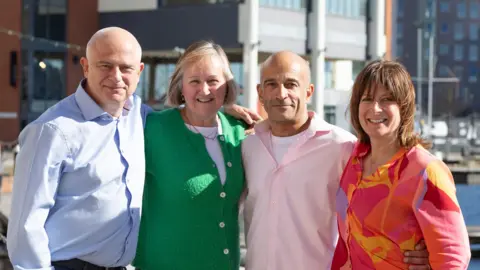 What Associates/Ipswich Book Festival Two men and two women stand together smiling at the camera. One man has a blue shirt on while the other has a pink shirt on with a white T-shirt underneath. One of the women has short blonde hair and wears a green cardigan with a white top underneath and the other woman has short brown hair and wears a red, pink, orange and yellow blouse. Ipswich Waterfront can be seen behind them. 