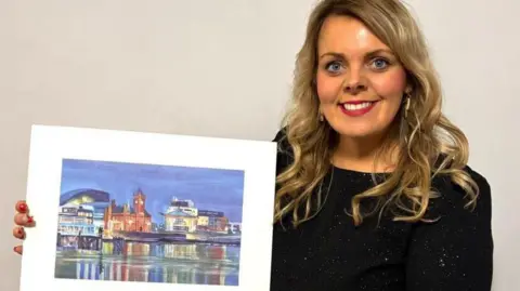 Getty Images A woman with long curly blojnd ehair wearing a black sparkly jumper and holding a painting of Cardiff Bay with the water in the front and colourful buildings behind.