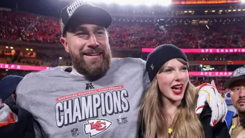 Getty Images A man with a grey sports t shirt and black baseball cap, with his arm round a woman with long blonde hair wearing a black beanie. They are stood in a stadium with crowds in red stands behind them.