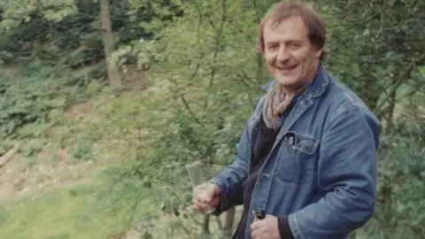 University of Leeds A photo of a man in a denim jacket wearing a scarf and holding a bottle of champagne and a glass. 