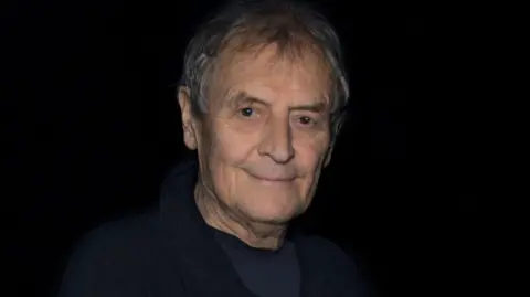 Getty Images A man with grey hair wearing a navy blue top and jacket, against a black background. 