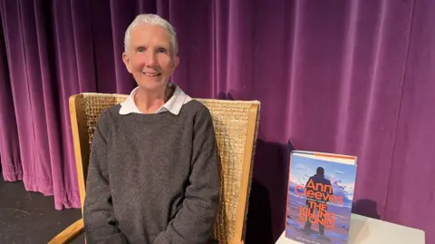 BBC Author Ann Cleeves, who has short white hair sits smiling in a woven wooden chair. She is wearing a dark grey sweater over a white collared shirt. Behind her is a stage backdrop of deep purple curtains. Next to her is her new book, The Killing Stones which is standing on a small white table. 
