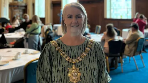 Tracy Kelly is wearing a green and black shirt. She has a golden chain around her neck, it has a large golden emblem on it. She has short blonde hair. Behind her is a busy room with people sitting at round tables on chairs. The room has large windows. It also has a blue carpet. 