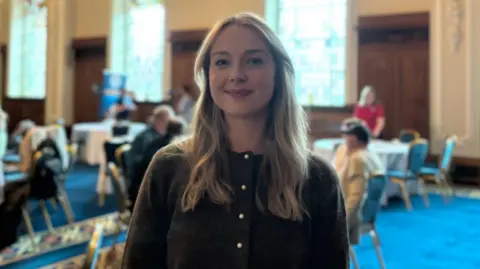 Megan Fletcher is wearing a brown top with silver buttons. She has long blonde hair down to below her shoulders. Behind her is a busy room with people sitting at round tables on chairs. The room has large windows. It also has a blue carpet. 