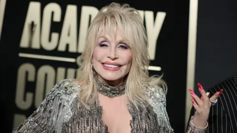 Getty Images Dolly Parton is wearing a silver dress. She has medium length blonde hair to her shoulders. She has long pink nails on her hand. She also has a silver necklace on. The background behind her is a billboard with white writing on a black background. 