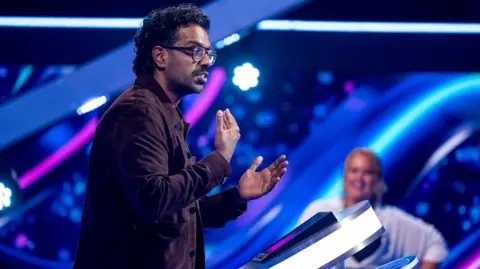 Romesh Ranganathan filming an episode of The Weakest Link, standing behind the presenter's podium in the BBC studio