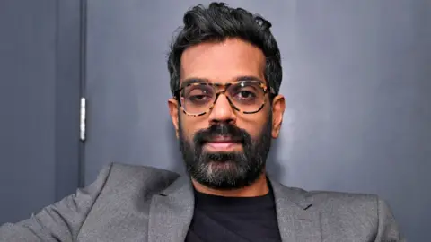 BBC Romesh Ranganathan pictured in front of a navy blue wall, looking at the camera, in March 2024