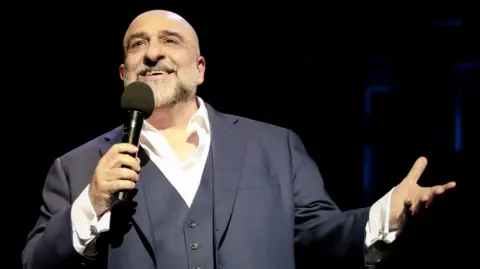 Getty Images Omid Djalili pictured performing on stage in 2023 in a grey suit, against a black backdrop