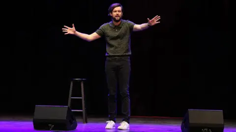 Jeremychanphotography/Getty Images A picture of comedian Jack Whitehall performing on stage. He is wearing a polo shirt, black jeans and white trousers and has his arms both out raised.