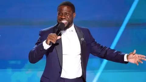 Getty Images A picture of Kevin Hart speaking into a mic, in a dark blue suit against a blue backdrop