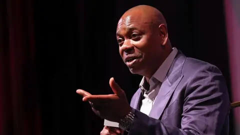 Getty Images Dave Chappelle speaks at the premiere of "Dave Chappelle: Live In Real Life" during the 2025 Martha's Vineyard African American Film Festival at Martha's Vineyard Performing Arts Center on August 08, 2025 in Massachusetts. He is wearing a purple suit and holding a microphone.