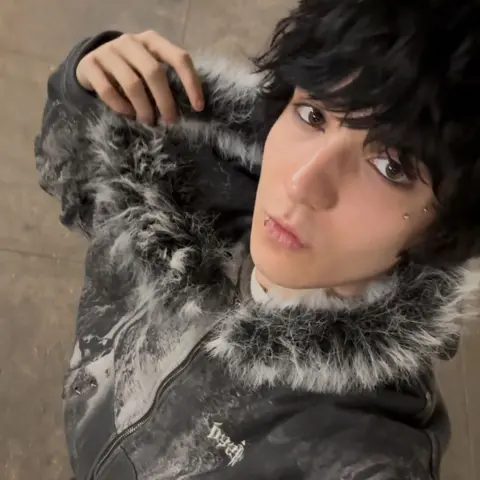 Ifesinachi Mamah Ifesinachi Mamah, taking a selfie wearing a grey coat with a fluffy trim hood. They have black hair and a lip and face piercings.