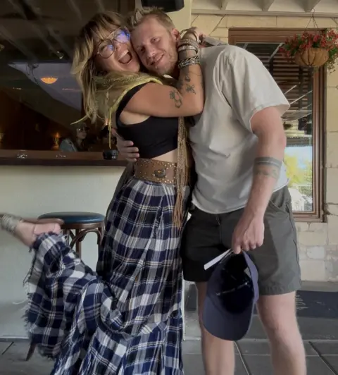 Jessica Raialo Jessica, wearing a long blue checked skirt and black sleeveless top, hugs her partner who is wearing dark grey shorts and a light t-shirt and holding a blue cap.