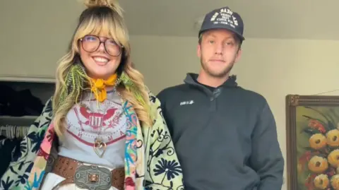 Jessica Raialo Jessica Raialo wearing a green and blue flower patterned jacket, orange neck scarf, grey and red t-shirt and a belt, next to her boyfriend wearing a dark cap and long-sleeved top. 