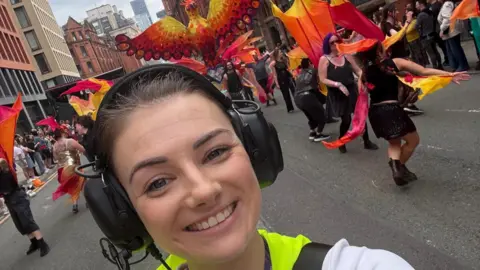 Abbie Ashall Abbie is close to the camera, smiling. Her dark brown hair is tied back and she is wearing large black headphones. Behind her down the street is the parade. You can see dancers wearing black with orange, pink and yellow streamers. 