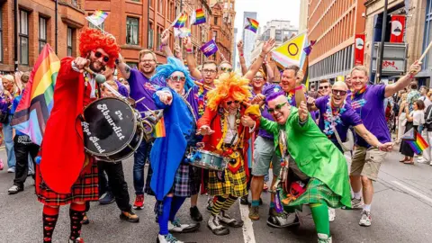 Getty Images Manchester Pride parade. People dressed in brightly-coloured suits holding pride flags. Some are holding large drums and colourful kilts. They are posing with their arms held out and smiling in the street. 