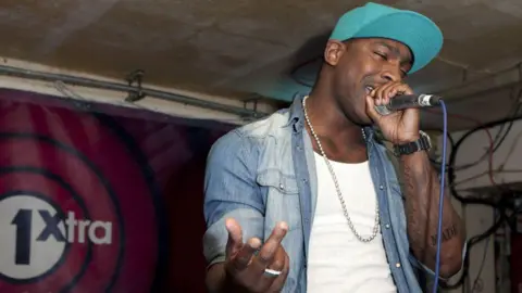 John Rahim/ Shutterstock A young Skepta, wearing an open denim shirt over a white t-shirt, rapping into a microphone in the BBC Radio one extra studios. 