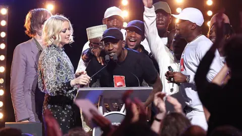 Tim P. Whitby/Getty Images Skepta, wearing a black t-shirt and black cap, on stage accepting the Mercury Prize. He is surrounded by friends and family