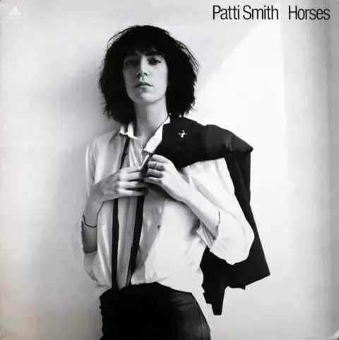 Patti Smith/Robert Mapplethorpe Foundation Patti Smith in white shirt with a dark ribbon hanging around her collar, and holding a dark jacket over her shoulder