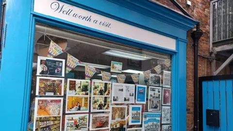 Angela O'Connor A shop window framed in bright blue paint with the words ‘Well worth a visit’ displayed at the top. The window features a collage of photographs, articles, and printed materials showcasing the store’s history and events.
