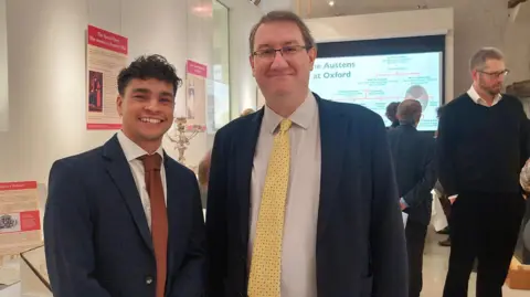 Exhibition curators Dr Timothy Manningmore and Michael Riordan, archivist at St John's College and The Queen's College, posing for a picture during the exhibition preview. Various artefacts could be seen behind them. A panel illustrating the Austen family tree and Oxford connections is displayed on a panel. 