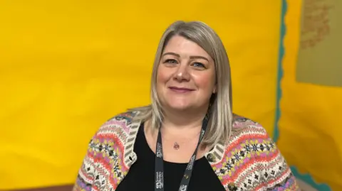 A blonde haired woman with a colourful cardigan and black top smiles at the camera