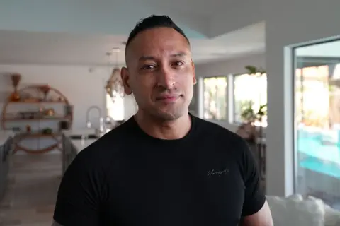 Tony Marquez stands in what appears to be a living room of a home. He is wearing a black T-shirt 