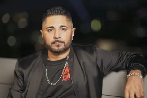 Jash Negandhi is seen sitting in a couch-like chair. He is wearing a black T-shirt and a black jacket. 