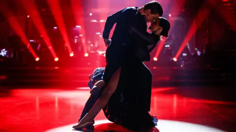 Nikita and Amber dancing on the strictly stage in a dark outfits. The lighting is red and the pair are holding each other in the centre of the stage. 