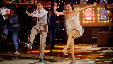 Amber and Nikita dancing on the strictly stage. They both have their arms and one leg in the arm in a coordinated move. 