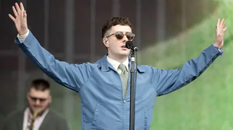PA Media Joesef performing onstage at an outdoor music festival - he is standing onstage and singing, while wearing a shirt, tie and blue jacket