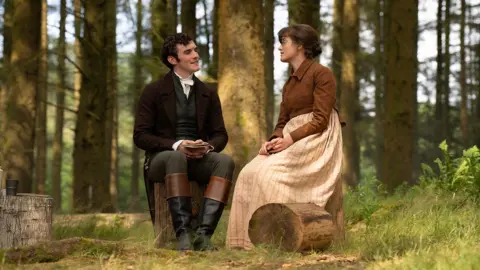 BBC/Bad Wolf/James Pardon Left to right: Mr Hayward (Dónal Finn) wearing a suit in a forest with Mary Bennet (Ella Bruccoleri), who is dressed in a beige and brown dress.